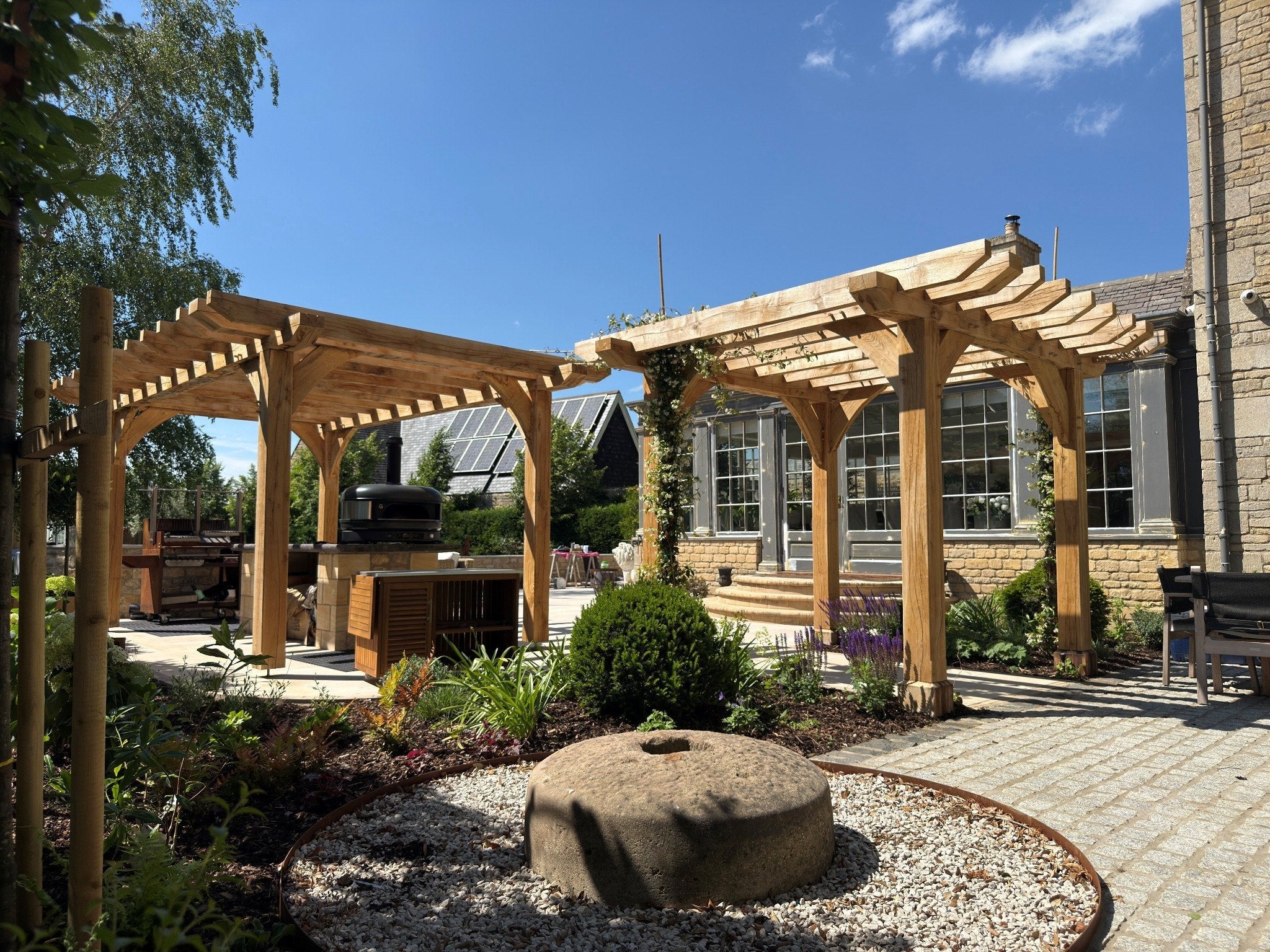 Two traditional oak pergolas over a garden patio and outdoor kitchen beside a stone house