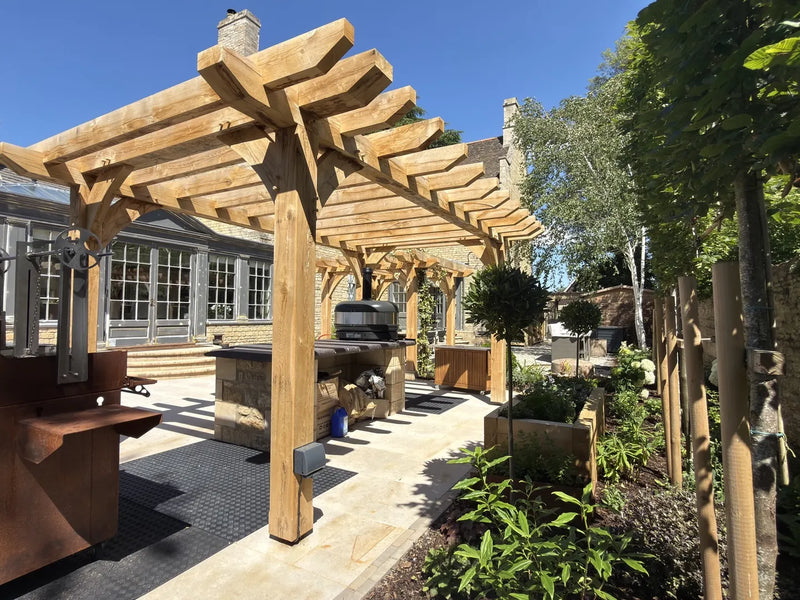 Oak pergola with curved braces over an outdoor kitchen beside a stone house